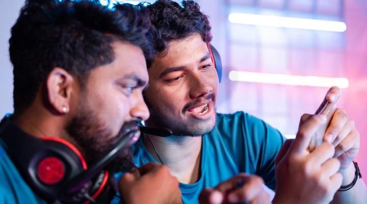 Two men wearing gaming headsets watch something on a smartphone.